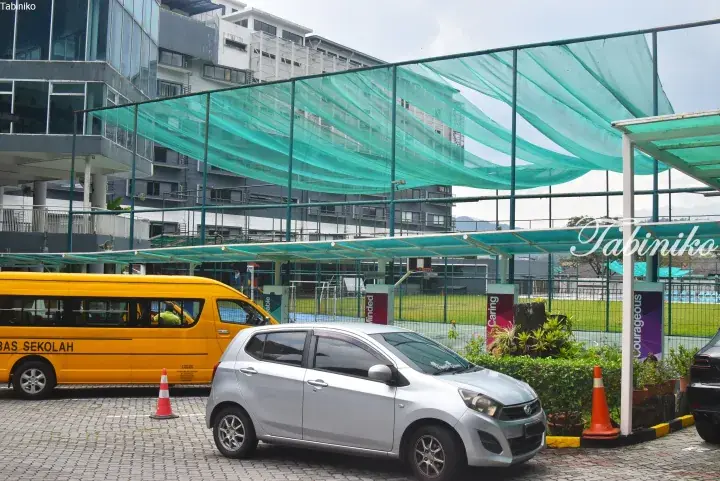Yellow school bus and silver car parked outside a modern school building with green netting over a sports court in Malaysia.
