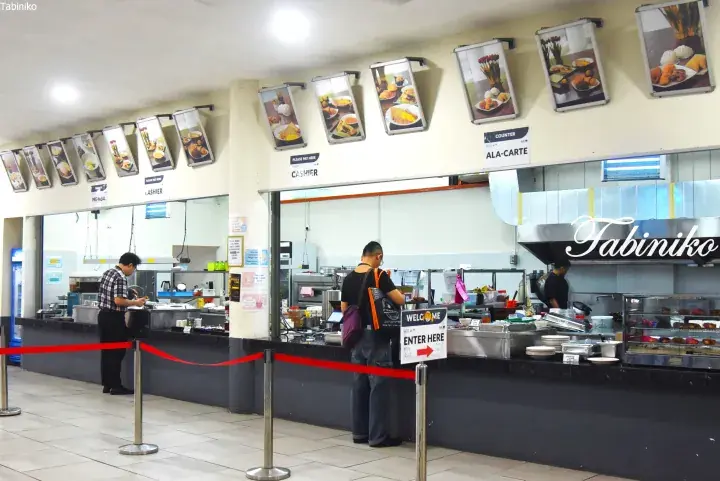 Customers ordering at the counter of a Malaysian food court with menu displays and a ‘Welcome Enter Here’ sign.