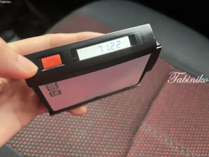 Hand holding a black digital luggage scale displaying 7.122 kg, with a red button and pink surface visible in the background.