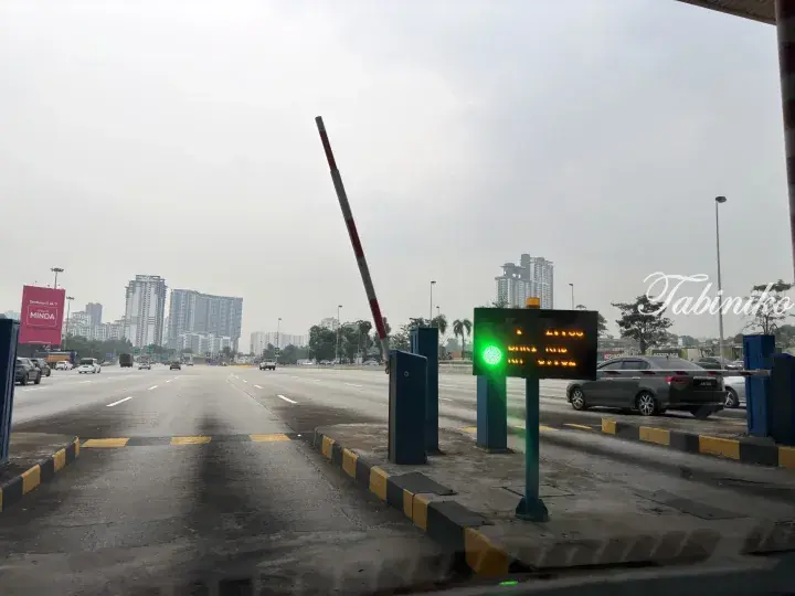 Toll booth with green light and raised barrier on a highway, with city skyline and high-rise buildings in the background.
