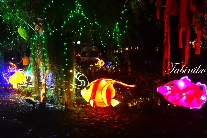 Colorful illuminated fish lanterns and green fairy lights decorate trees at a night festival in Malaysia.