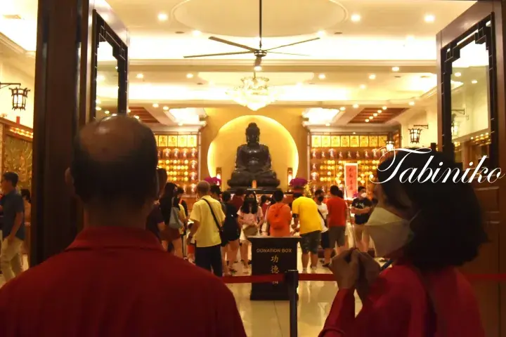 Devotees pray before a large Buddha statue inside a brightly lit temple hall with donation boxes.