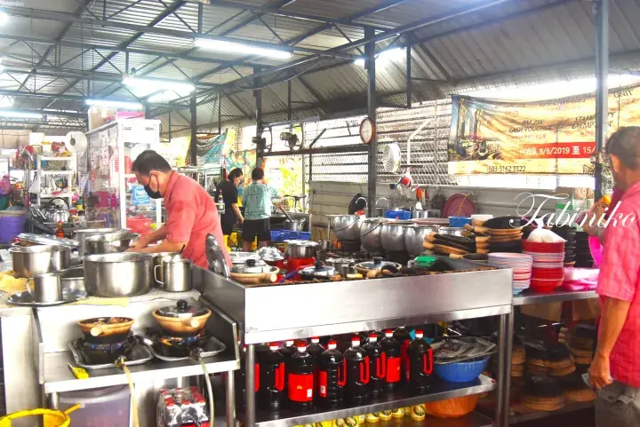 Chef cooking in a busy open-air Malaysian kitchen with large pots and condiments.