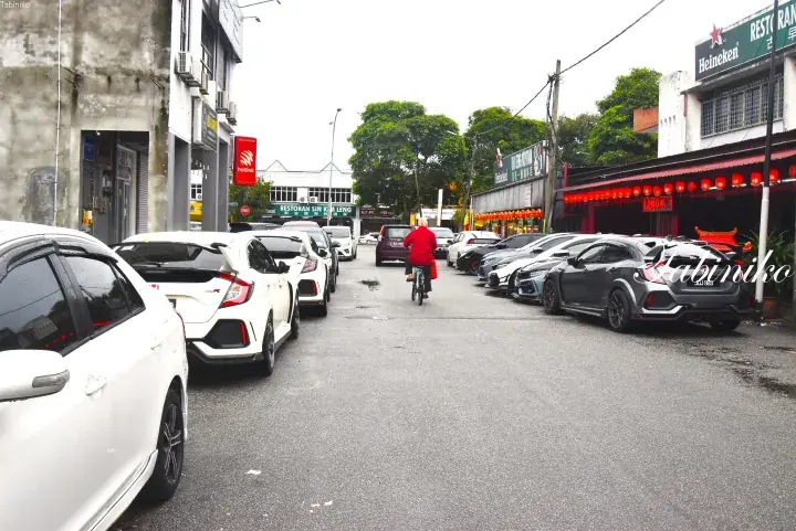 A street lined with parked Honda Civic Type R cars in Malaysia, with a cyclist riding past local shops and restaurants.