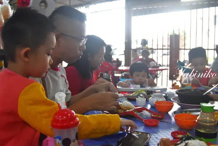 Family enjoying a meal at a Malaysian restaurant with children and adults sharing dishes at a blue table.