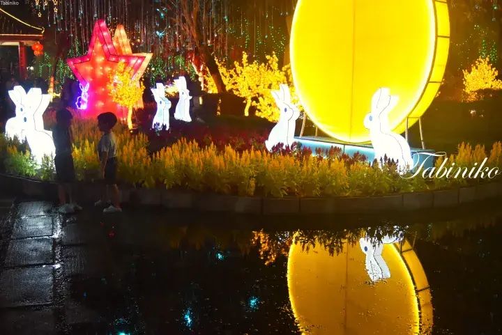 Illuminated rabbit lanterns and a giant yellow moon at a night festival, with reflections in the water.