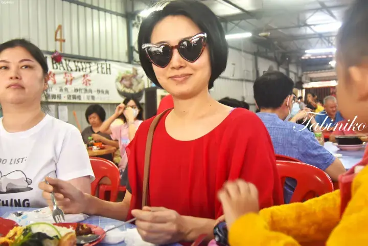 Woman in red top and heart-shaped sunglasses smiles at a table in a Malaysian hawker centre with a Bak Kut Teh sign.