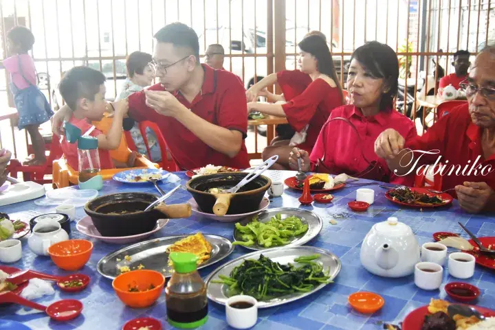Family enjoying a meal together at a Malaysian restaurant, featuring claypot dishes and vegetables on a blue table.