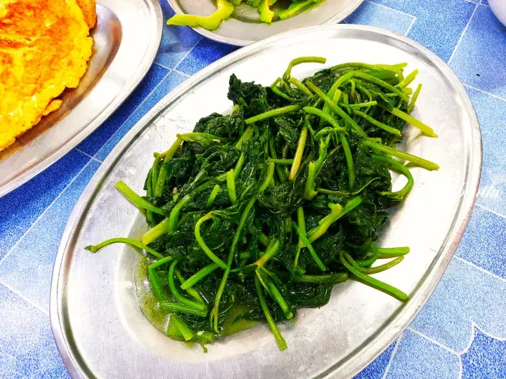Stir-fried spinach served on a metal plate alongside a golden omelette at a local Malaysian eatery.