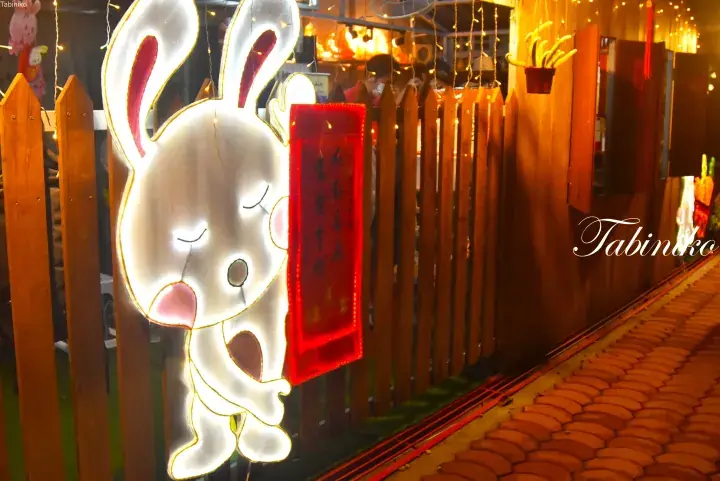 Illuminated rabbit lantern holding a red banner at a festive night market in Malaysia.