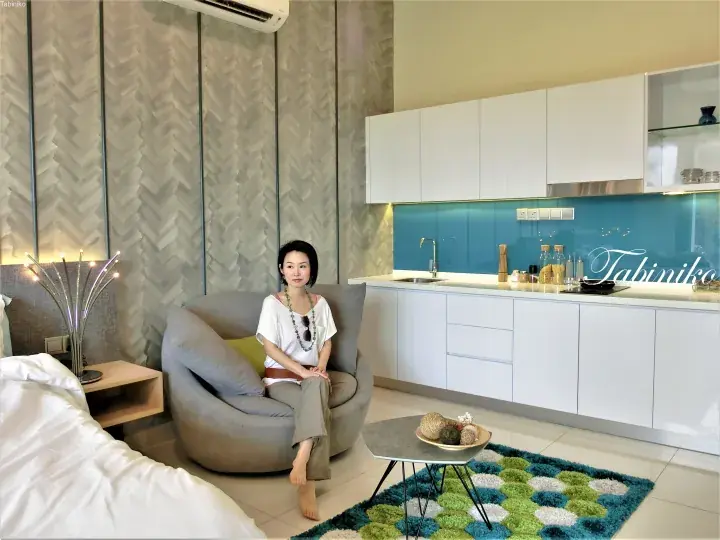 Woman sitting in a modern studio apartment with a kitchenette and textured wall panels.