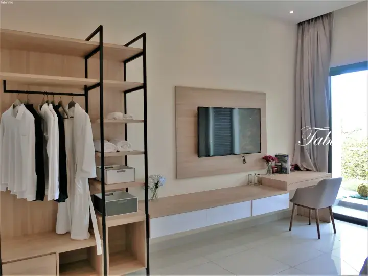 Modern hotel room interior featuring an open wardrobe with white robes and a wall-mounted TV above a floating desk.