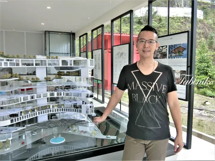 Man in black t-shirt stands beside a detailed architectural model of a high-rise building at a property showroom.