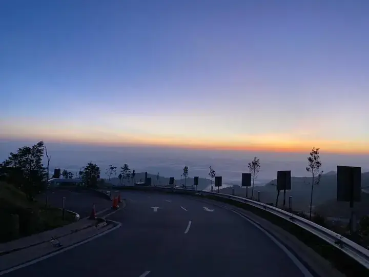 Winding mountain road at sunrise with sea of clouds below and colorful sky.