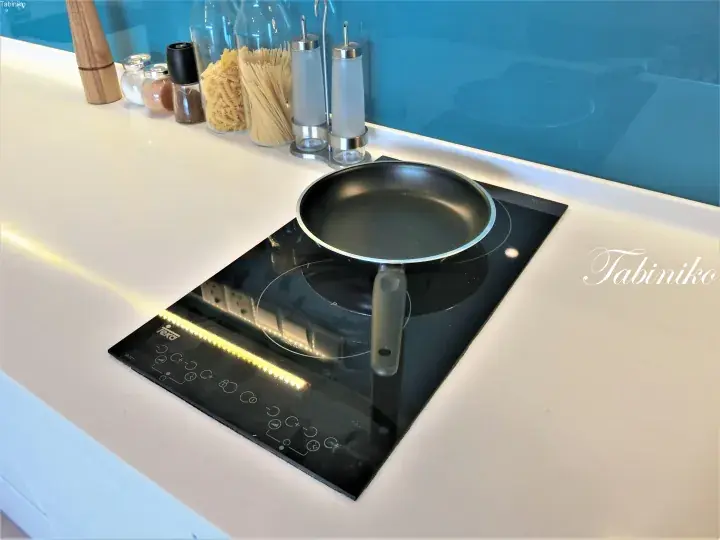 Modern kitchen featuring a black induction cooktop with a frying pan, white countertop, and blue glass backsplash.