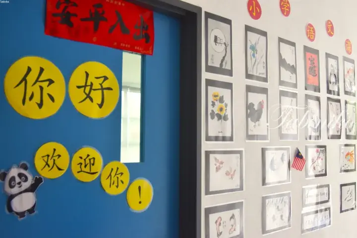 Blue classroom door with yellow ‘Hello’ and ‘Welcome’ signs, featuring a cartoon panda, next to a wall displaying student Chinese brush paintings.
