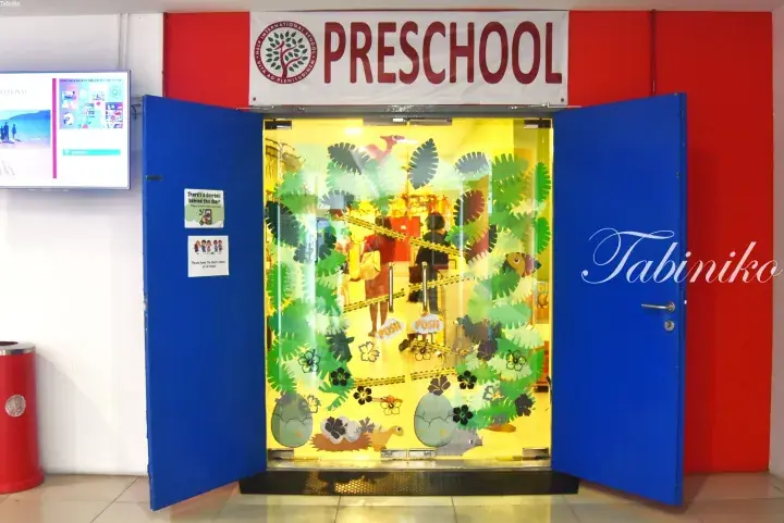 Entrance to HELP International School Preschool featuring blue doors, a red wall, and a glass door decorated with colorful paper leaves and animals.