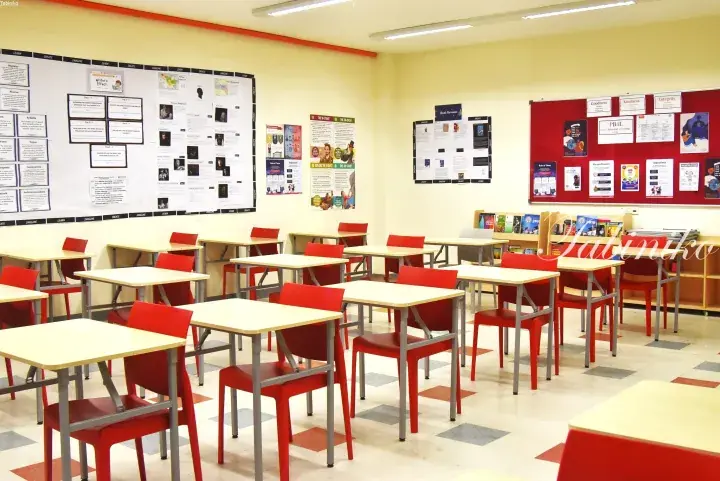 Empty classroom with red chairs and light wood desks, featuring educational posters and bulletin boards on the walls.