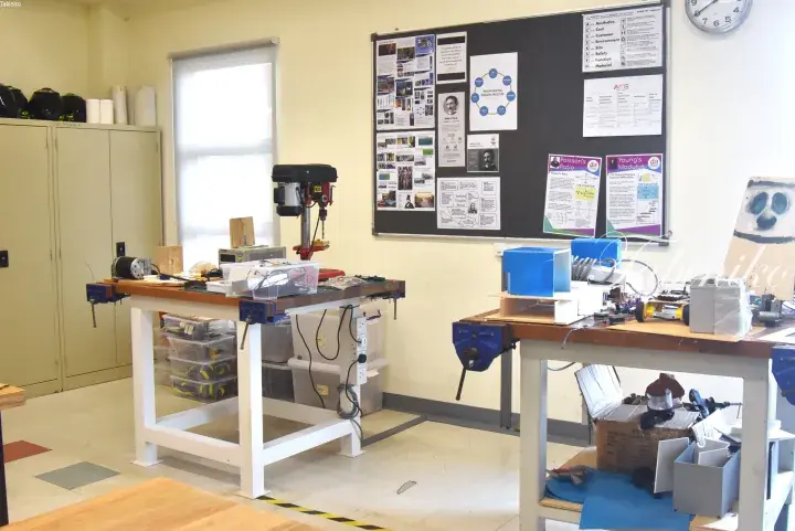 Workshop table with drill press and tools next to bulletin board with engineering posters in a classroom.