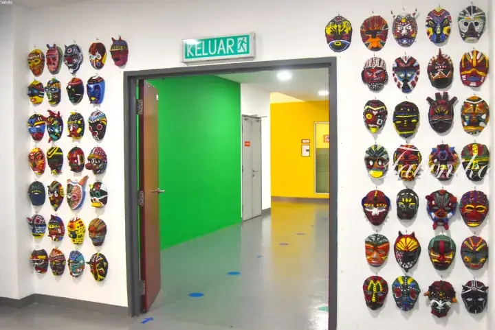 Colorful African masks adorn walls near a green screen room and exit door marked KELUAR.