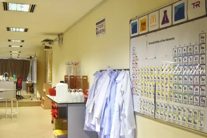 Science lab with periodic table poster, hanging lab coats, and glassware on a counter.