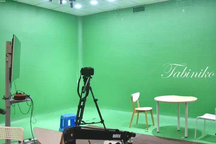 Green screen studio setup with camera on tripod, table, chairs, and RODE microphone for video production.