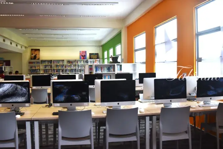 Modern library computer lab with rows of Apple iMacs, colorful orange and green walls, and bookshelves in the background.