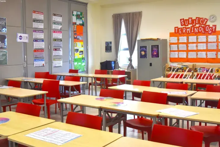 Empty classroom with red chairs, wooden desks, and educational posters on walls.