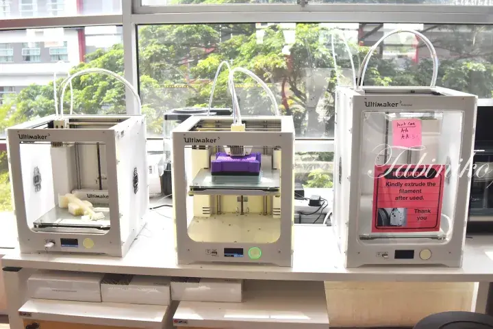 Three Ultimaker 3 Extended 3D printers on a table near a window with greenery outside.