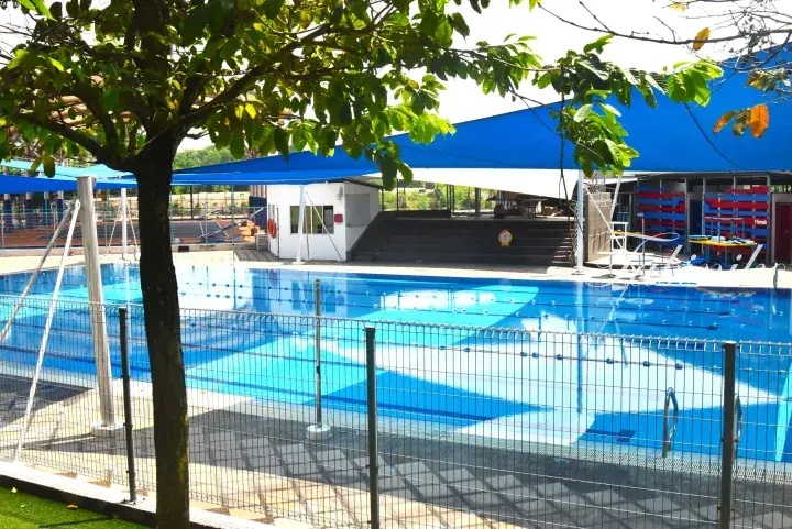 Outdoor swimming pool with blue lane dividers, shaded by blue canopies and surrounded by a metal fence and greenery.