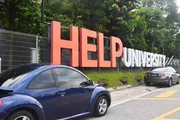 Large orange HELP University sign outside campus with parked cars and lush green trees in the background.