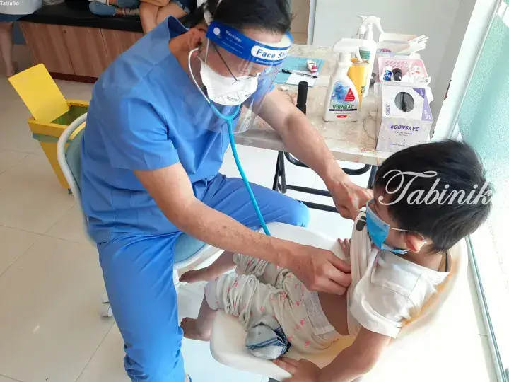 Doctor in blue scrubs and PPE examines a young child with a stethoscope at a clinic.