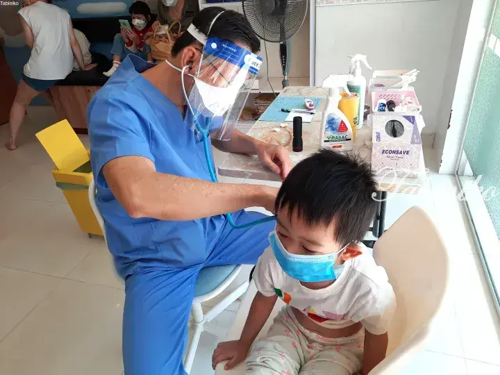 Doctor in blue scrubs examining a young child wearing a face mask in a clinic.
