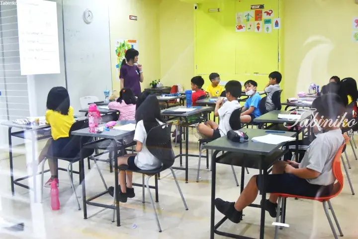 Teacher instructs students in a bright classroom with yellow walls and a whiteboard displaying English vocabulary.