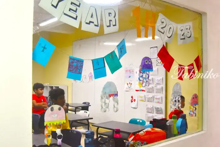 Classroom decorated with jellyfish crafts and a ‘YEAR 1H 2023’ banner, viewed through a window.