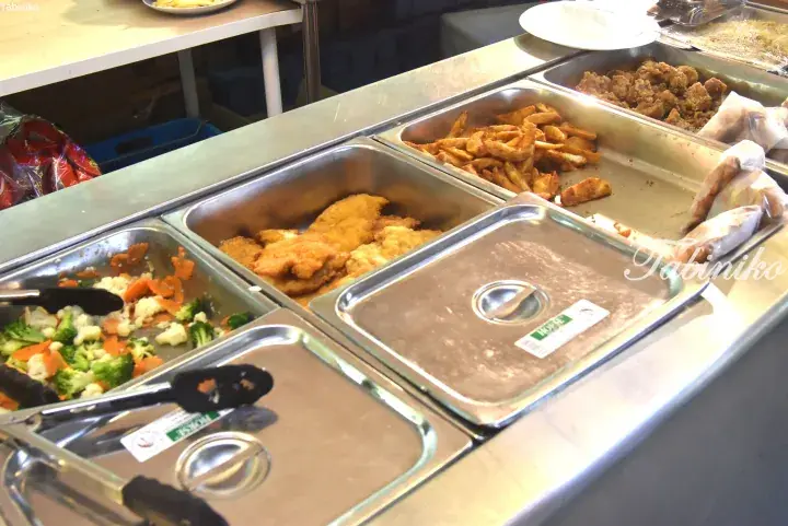 Buffet trays with fried chicken, wedges, and mixed vegetables at a Malaysian food stall.