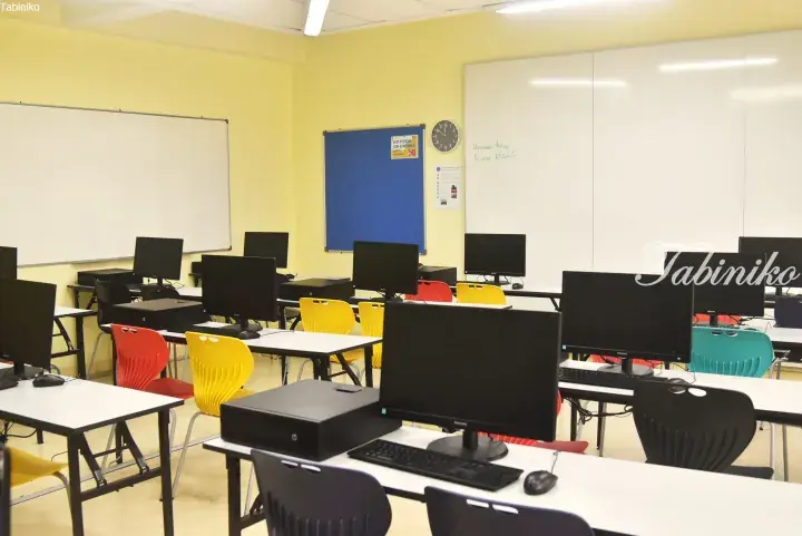 Bright computer lab with yellow walls, whiteboards, and colorful chairs arranged at desks with monitors.