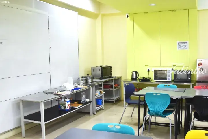 Bright communal kitchen with yellow walls, stainless steel tables, appliances, and colorful chairs in a Malaysia shared living space.