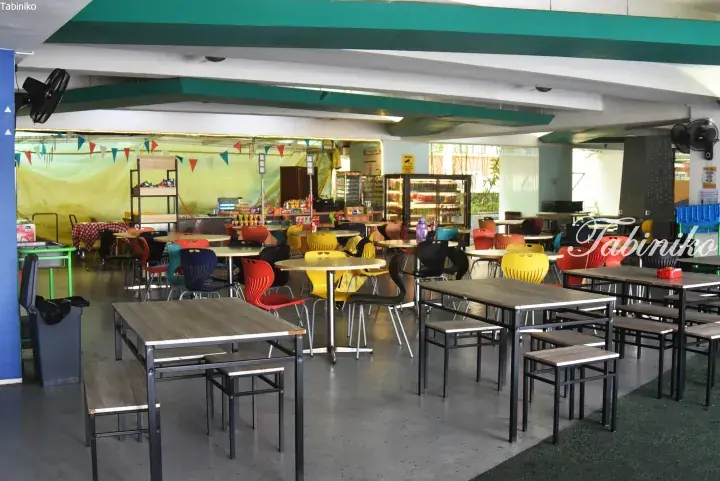 Colorful chairs and tables in a spacious, open-air school cafeteria with food stalls and festive bunting.