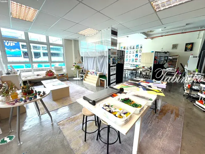 Bright art studio with tables of craft supplies, colorful student artwork, and large windows overlooking a pool area.