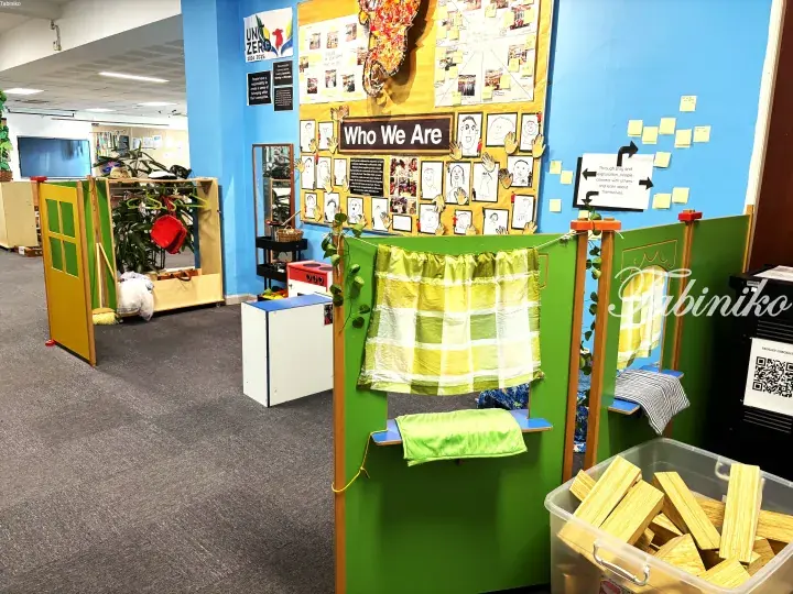 Colorful classroom play area with green wooden structures and a bulletin board titled &lsquo;Who We Are&rsquo; in a school.