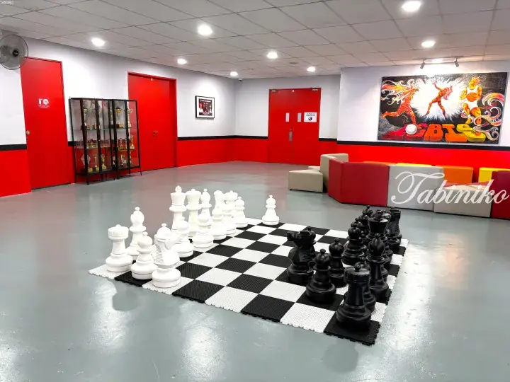 Giant chess set on a checkered floor in a modern lounge with red doors, trophy display, and vibrant sports mural.