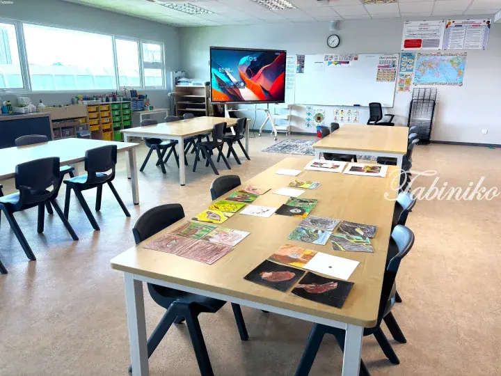 Bright art classroom with student paintings on wooden tables, smartboard, and storage bins.