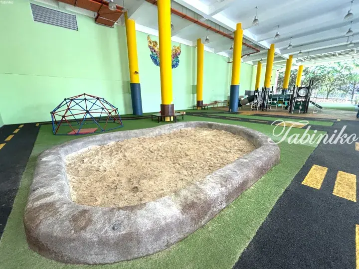 Indoor playground featuring a large sandpit, climbing dome, and play structures under a sheltered area with yellow pillars.