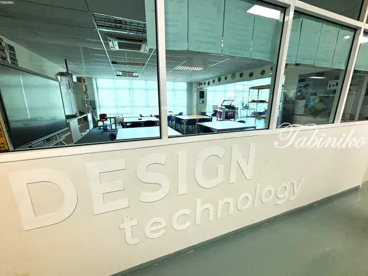 Glass-walled design technology classroom with white tables and chairs, viewed through a wall labeled DESIGN technology.