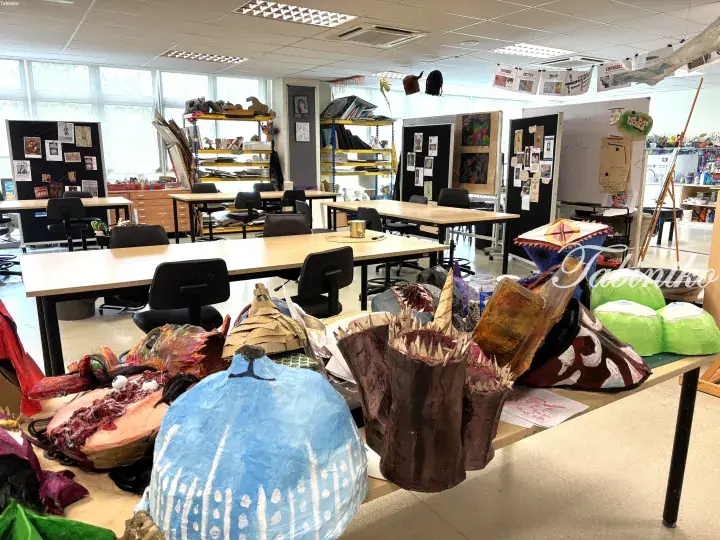 Colorful student art sculptures displayed on a table in a bright classroom with desks and shelves.
