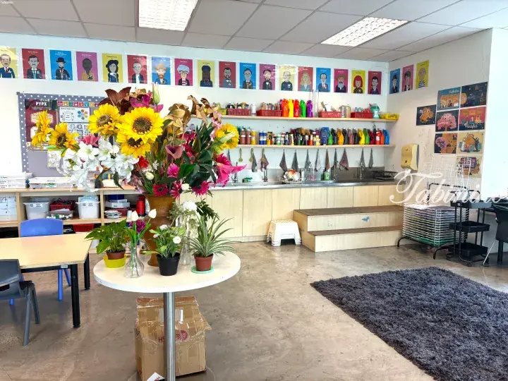 Bright classroom with large sunflower arrangement, artist portraits, and colorful paint supplies on shelves.
