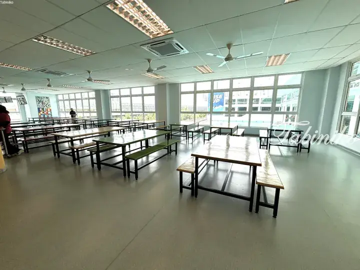 Spacious school cafeteria with long wooden tables, green benches, and large windows overlooking a swimming pool.