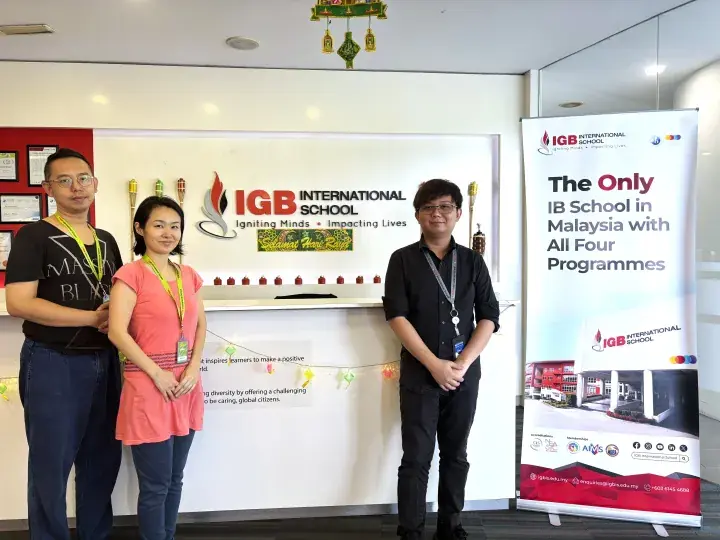 Three people standing at IGB International School reception with a banner highlighting its IB programmes.