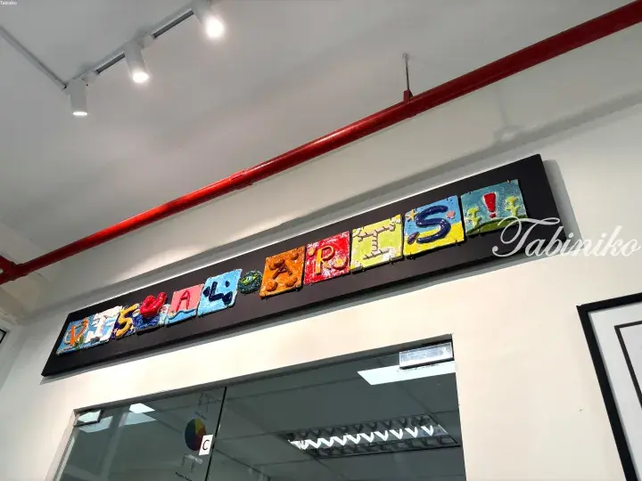 Colorful handcrafted sign spelling &lsquo;VISUAL ARTS&rsquo; mounted on a black board above a glass door.